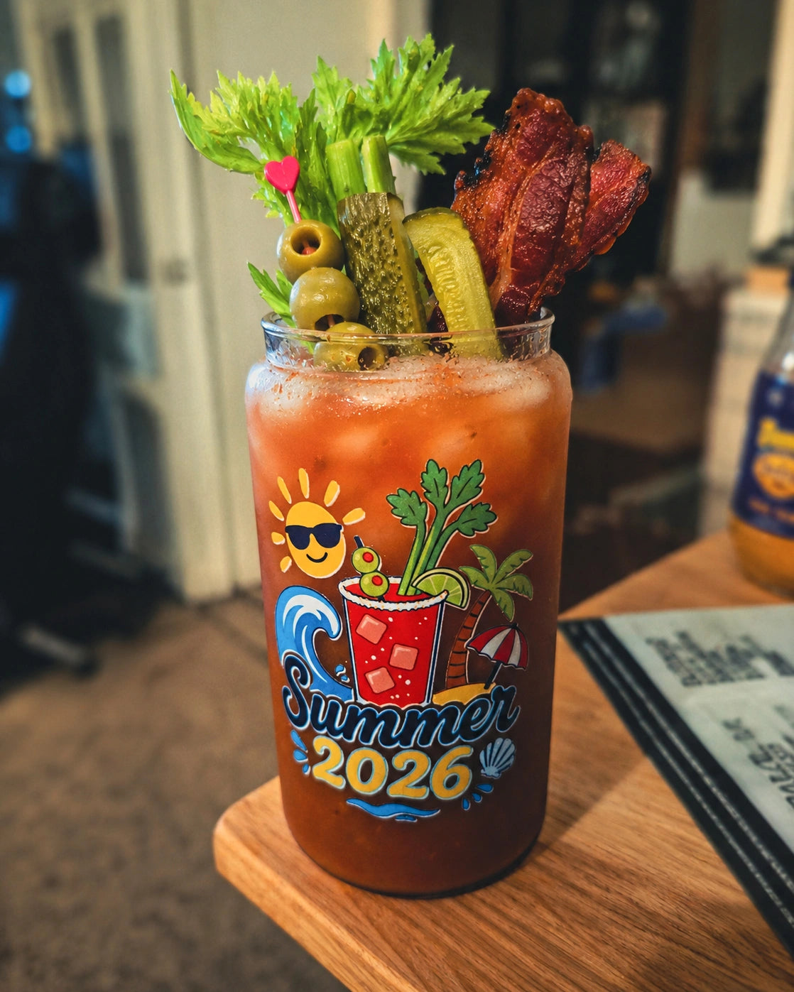 A Bloody Mary cocktail in a clear glass tumbler with a colorful 'Summer 2026' graphic, garnished with celery, pickles, olives, and a piece of bacon, sitting on a wooden table.