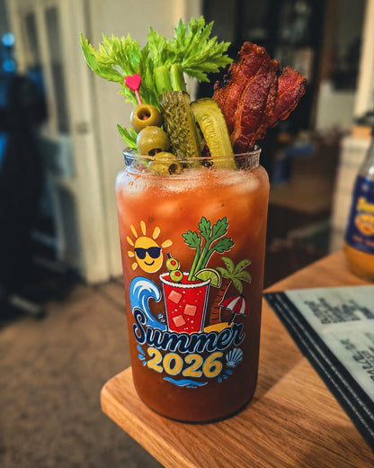 A Bloody Mary cocktail in a clear glass tumbler with a colorful 'Summer 2026' graphic, garnished with celery, pickles, olives, and a piece of bacon, sitting on a wooden table.